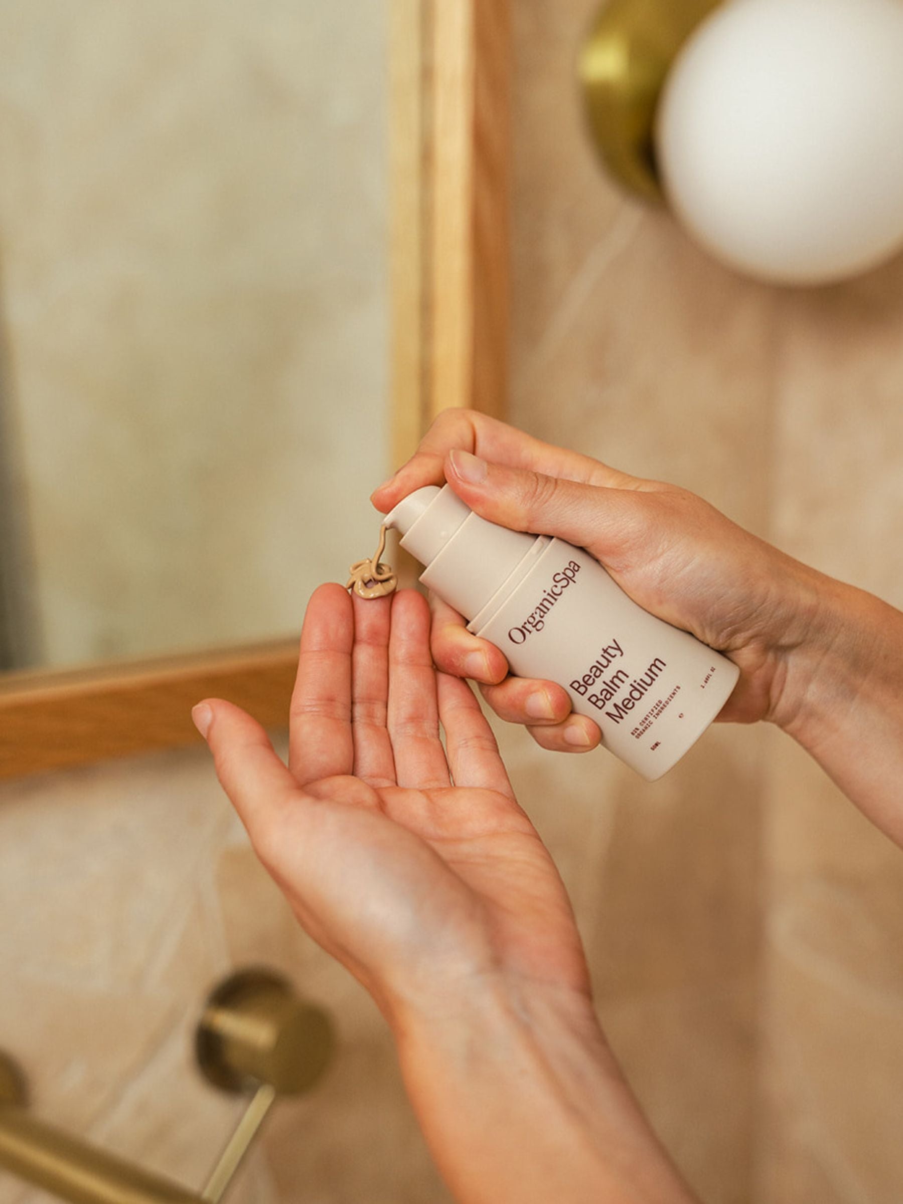 Hands dispensing Organicspa Beauty Balm Medium complexion cream into the palm in a bathroom, ready to apply as a tinted moisturiser.