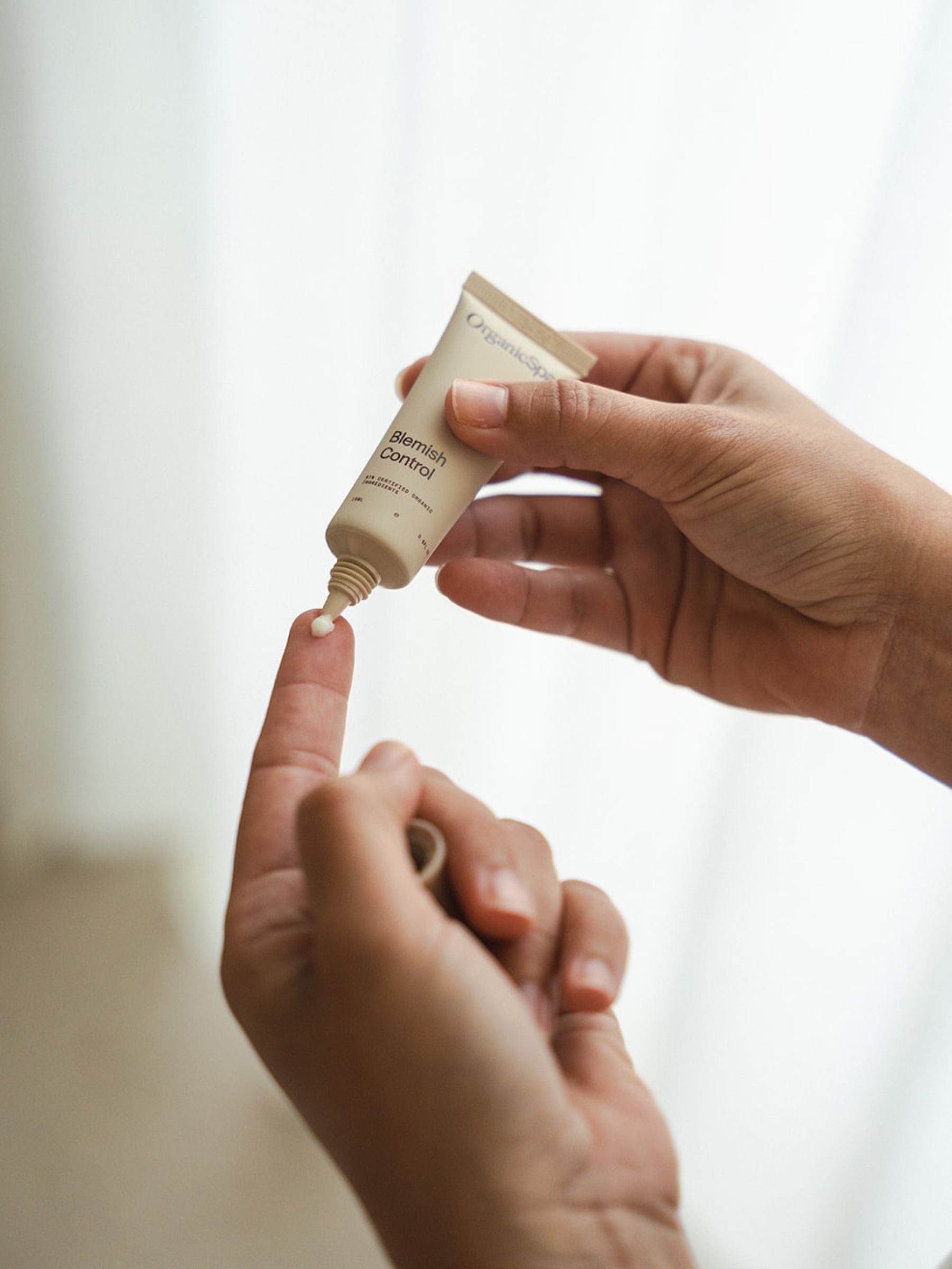 Close-up of hands squeezing a small amount of Organicspa Blemish Control spot treatment onto a fingertip, with a light curtain backdrop.