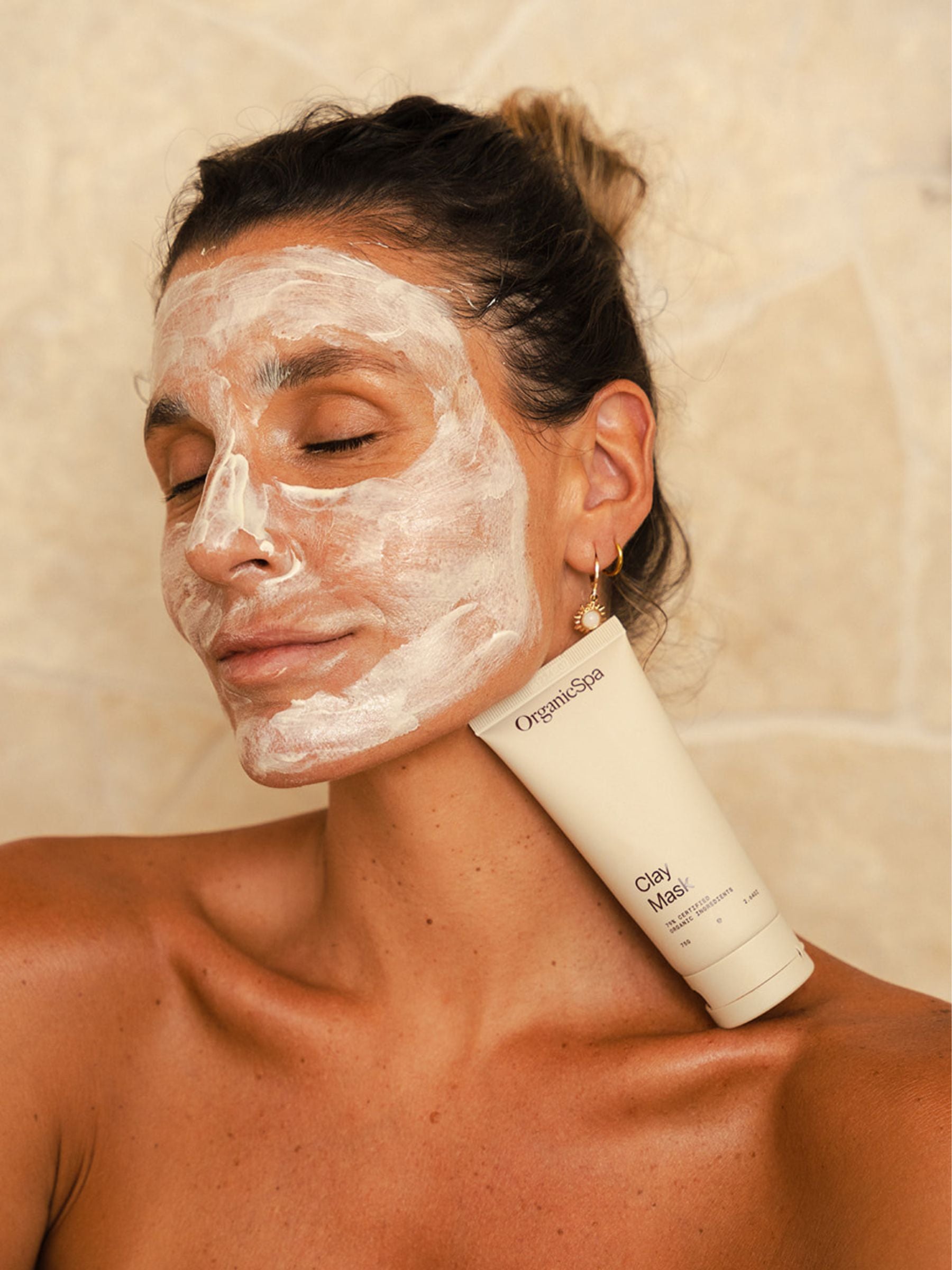 Woman with a full layer of Organicspa Clay Mask on her face, resting a tube of the mask on her shoulder against a tiled bathroom wall.