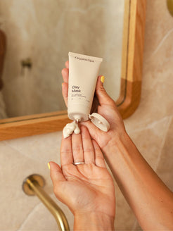 Hands squeezing a creamy amount of Organicspa Clay Mask from the tube into the palm in front of a bathroom mirror and brass tap.