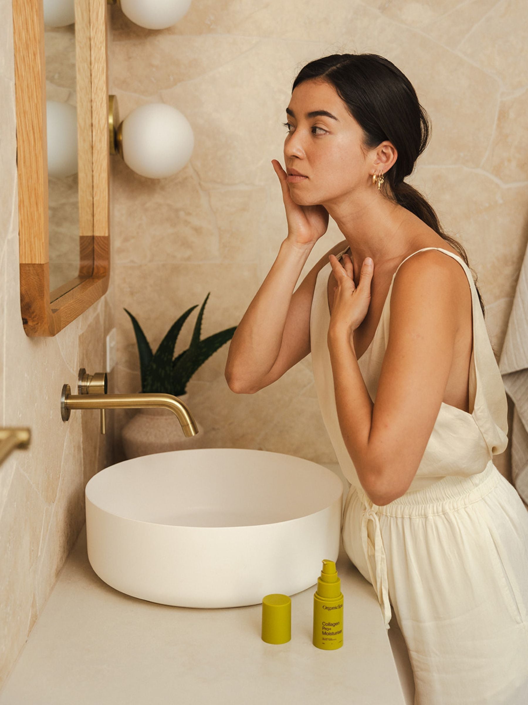 Woman standing at a bathroom basin applying Organicsceuticals Collagen Pro+ Moisturiser to her face, with the yellow bottle sitting beside the sink.
