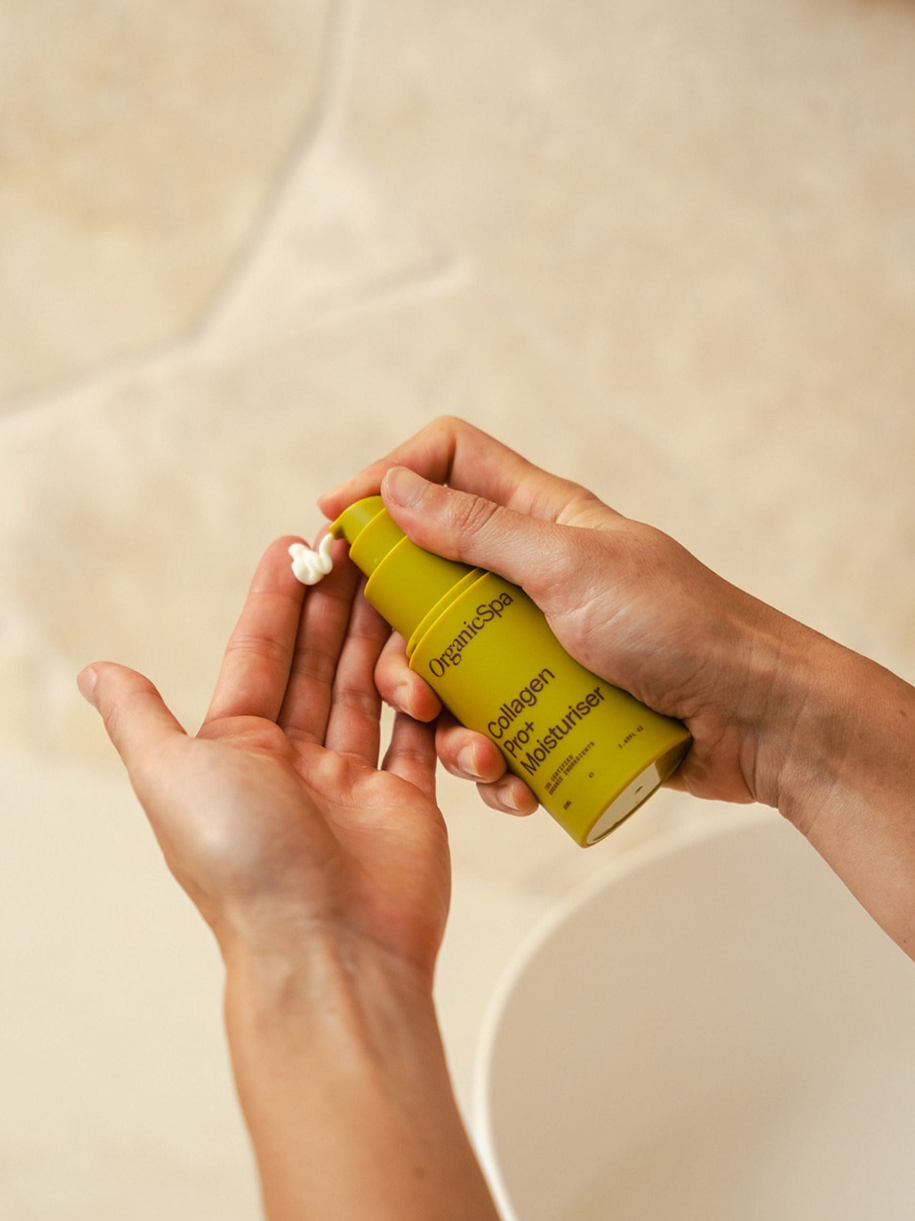 Hands dispensing Organicsceuticals Collagen Pro+ Moisturiser from a yellow pump bottle, showing the creamy certified organic formula before application.