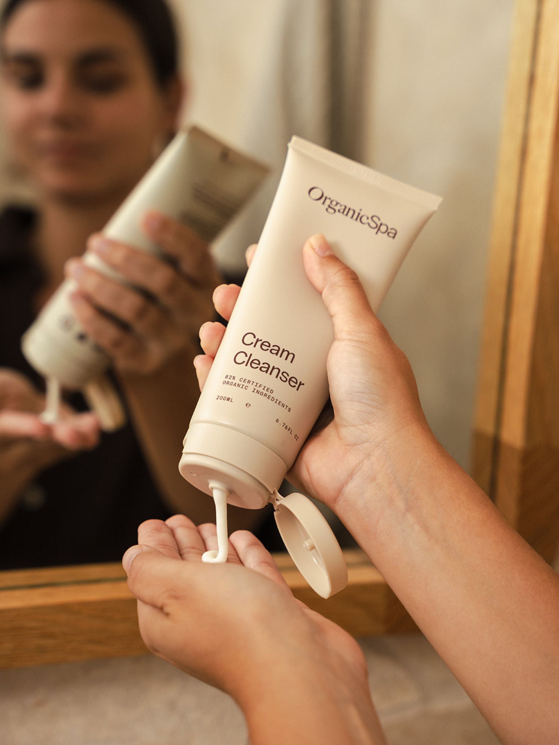 Person squeezing Organicspa Cream Cleanser from a tube into their hand in front of a bathroom mirror, showing the gentle certified organic cream cleanser in use.