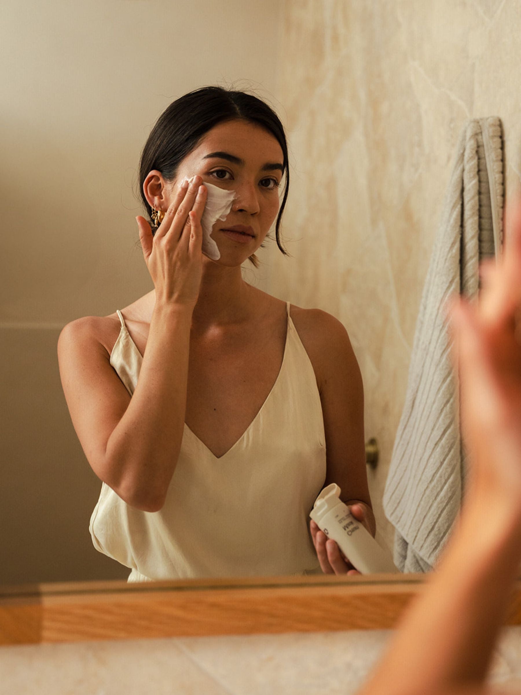Woman in a cream camisole applying Organicspa Cream Mask to her cheek while looking into a bathroom mirror, holding the tube in her other hand.