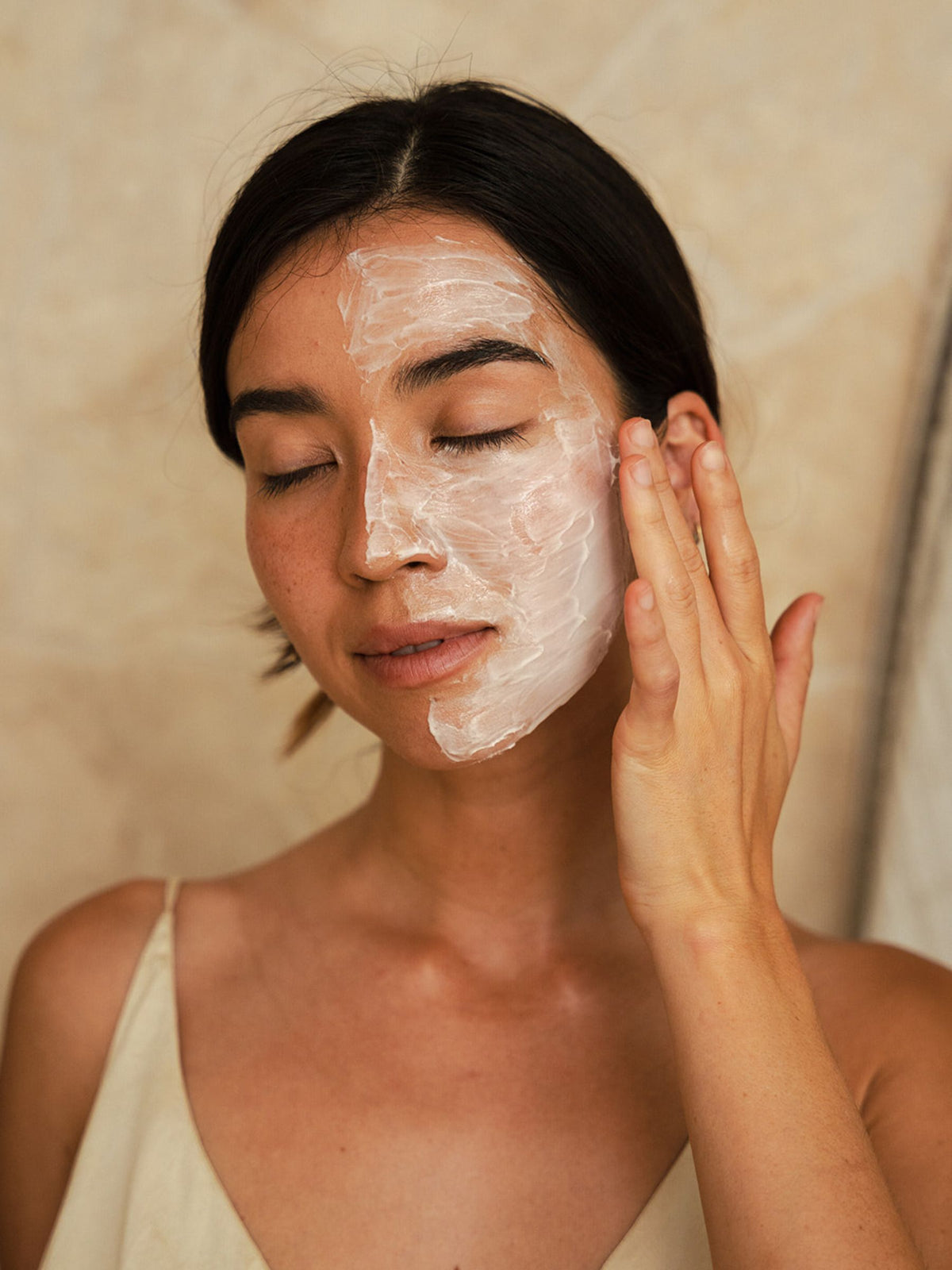 Close-up of a woman with eyes closed, smoothing a generous layer of Organicspa Cream Mask over one side of her face.