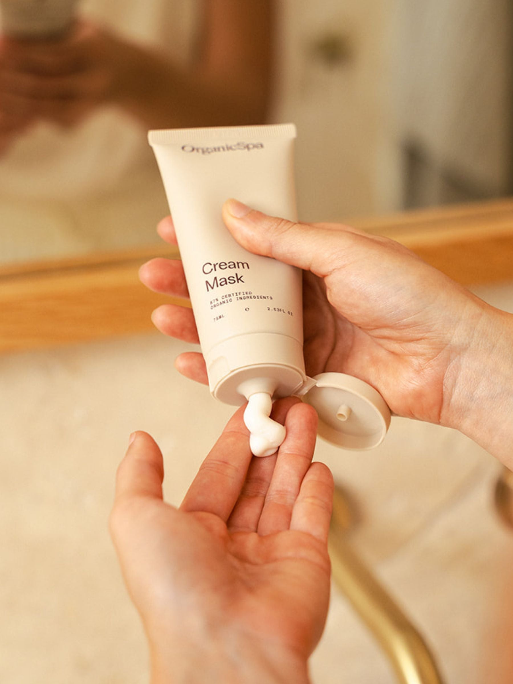 Hands holding a tube of Organicspa Cream Mask, squeezing a swirl of creamy mask onto fingertips in a bathroom setting.