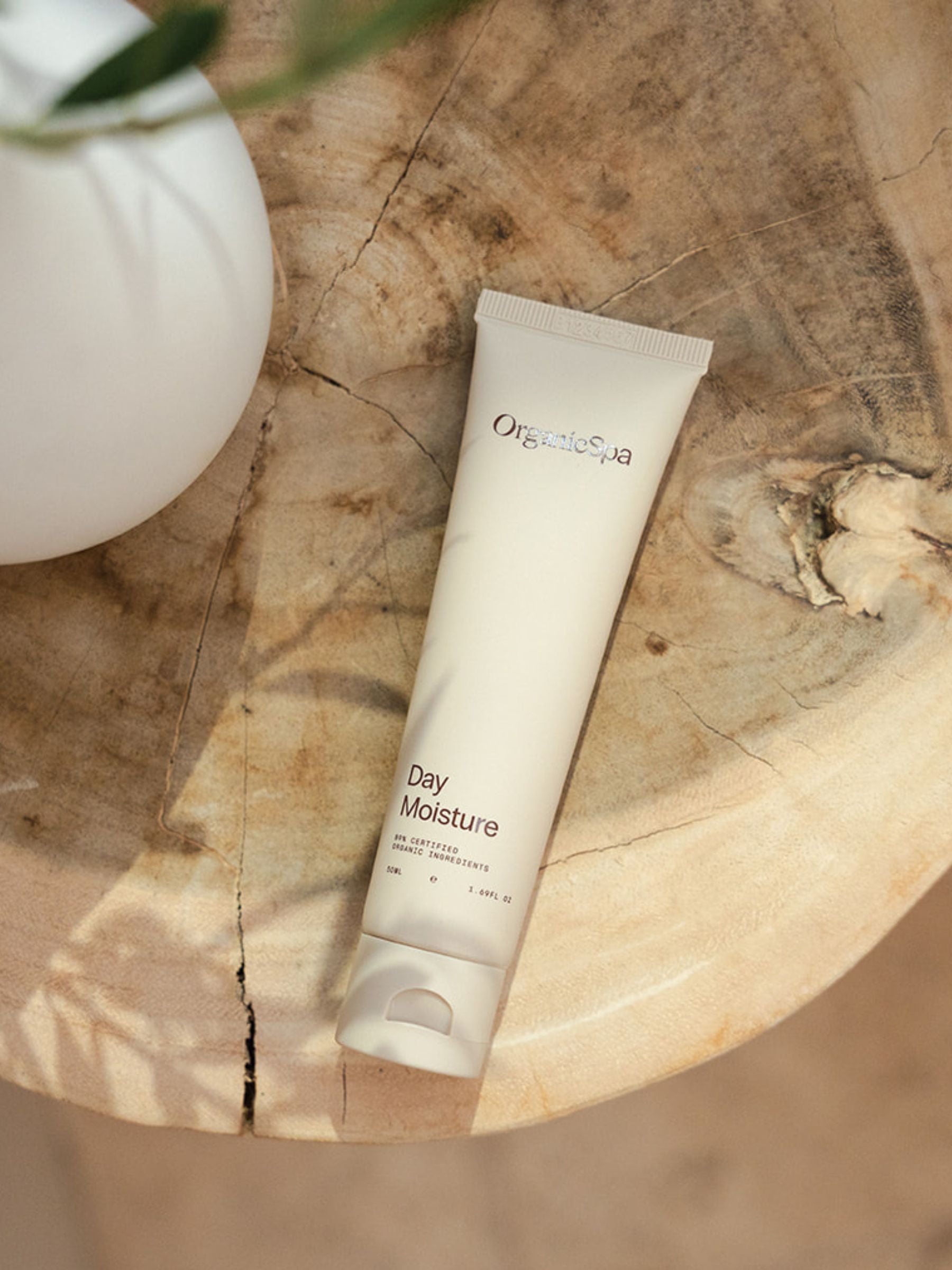 Tube of Organicspa Day Moisture facial moisturiser styled on a round wooden side table beside a white vase in soft natural light.