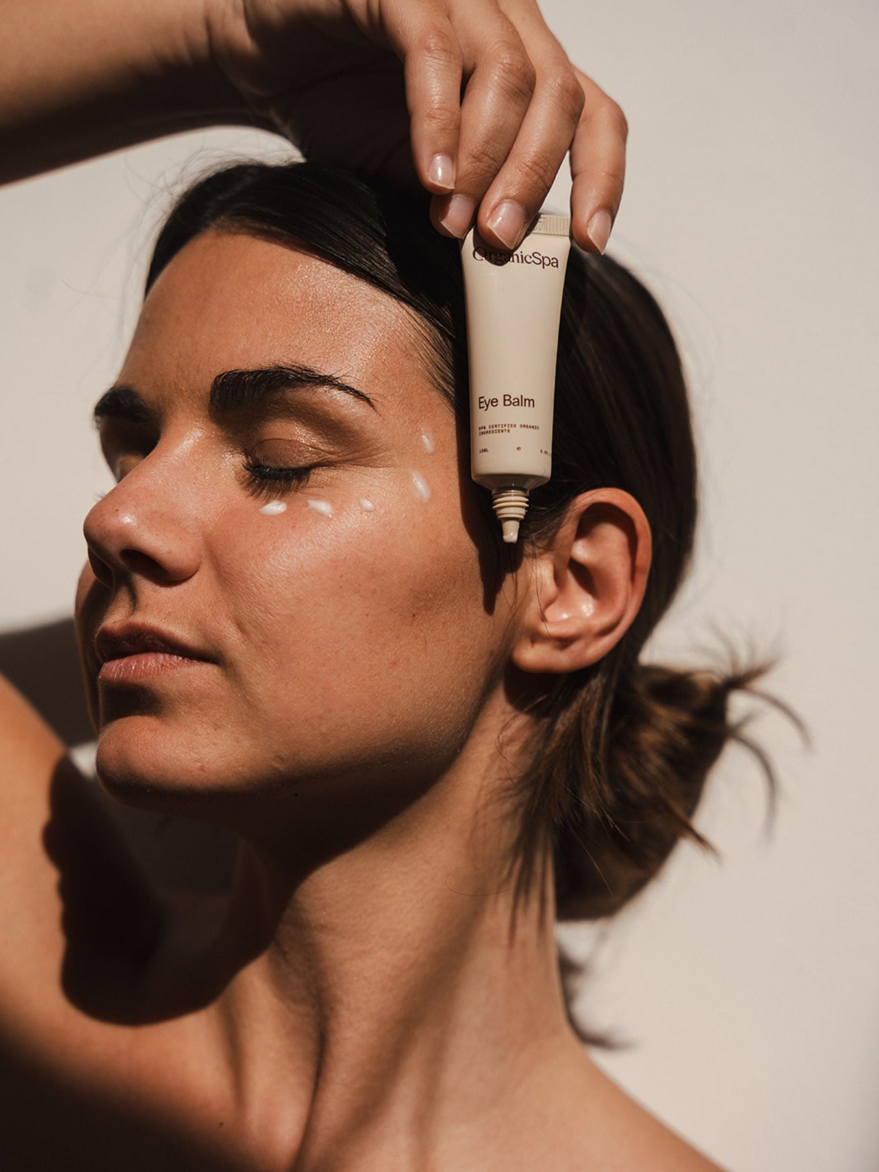 Woman in warm natural sunlight with eyes closed, holding Organicspa Eye Balm above her temple as small dots of product sit under her eye, ready to be gently blended in.
