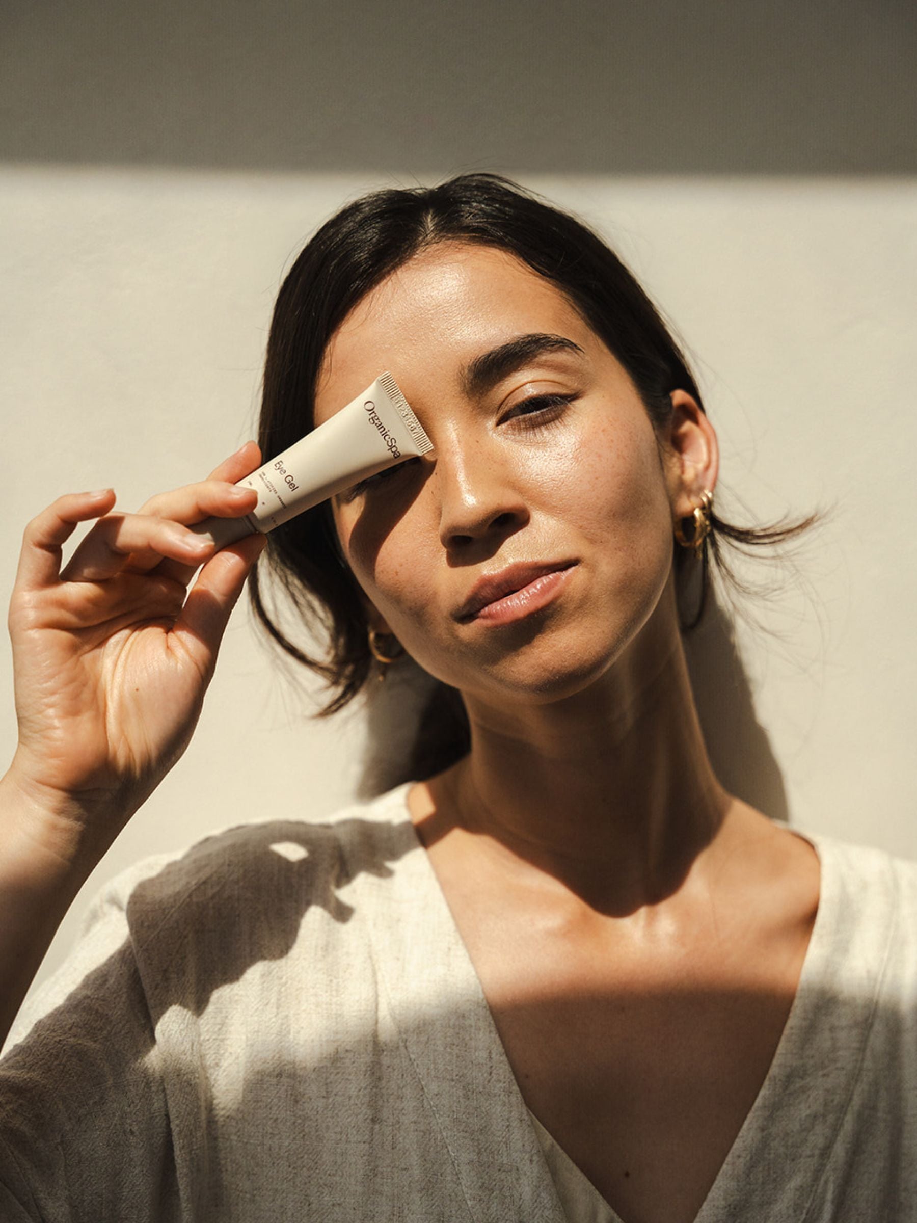 Woman holding Organicspa Eye Gel tube beside her eye in warm natural sunlight, showing a simple certified organic eye care ritual.