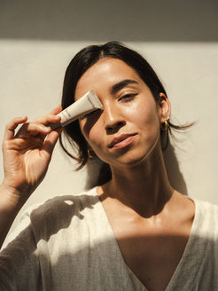 Woman holding Organicspa Eye Gel tube beside her eye in warm natural sunlight, showing a simple certified organic eye care ritual.