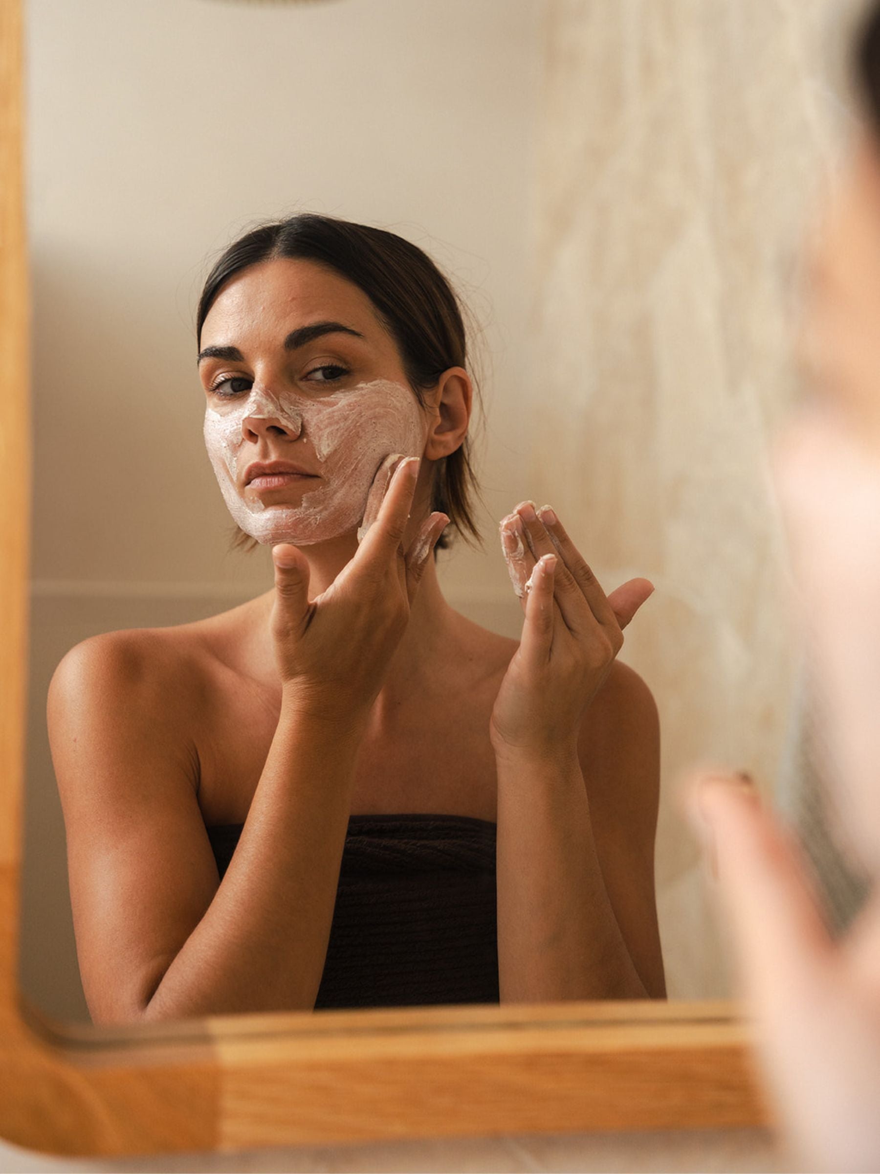 Woman applying Organicspa Face Exfoliant in front of a bathroom mirror, creamy certified organic exfoliant visible on her face.