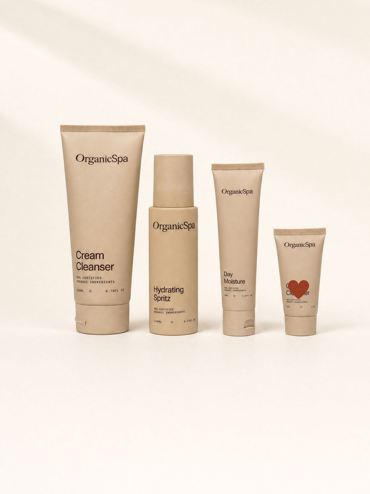 The four products in the OrganicSpa Hydrate + Replenish Collection lined up in sand-coloured packaging: Cream Cleanser 200ml at 80% Certified Organic Ingredients, Hydrating Spritz 140ml at 96% Certified Organic Ingredients, Day Moisture 50ml at 80% Certified Organic Ingredients, and a 30g travel size Cream Cleanser marked with a red heart.