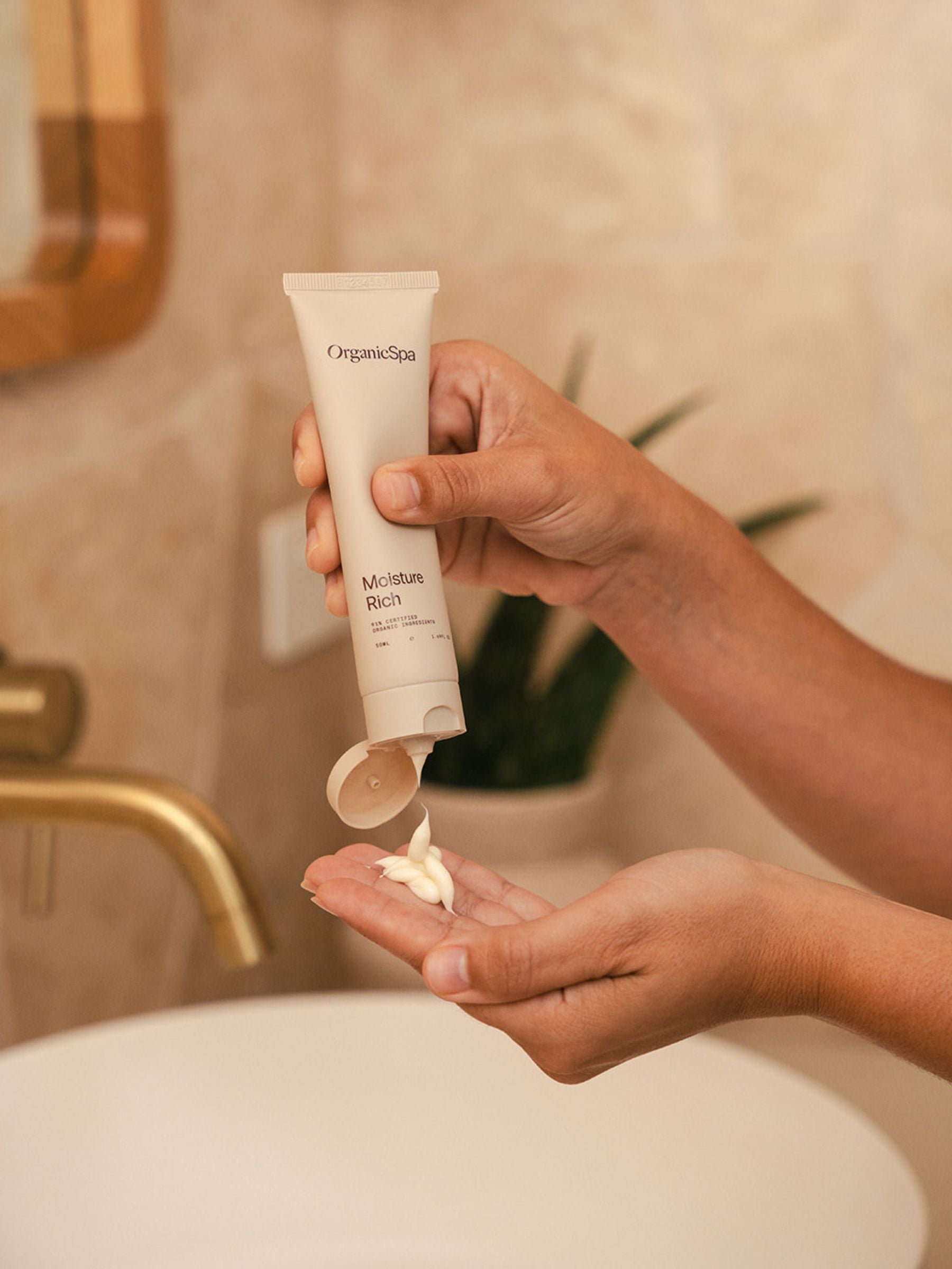 Hands holding an Organicspa Moisture Rich tube and dispensing moisturiser into the palm above a bathroom basin.