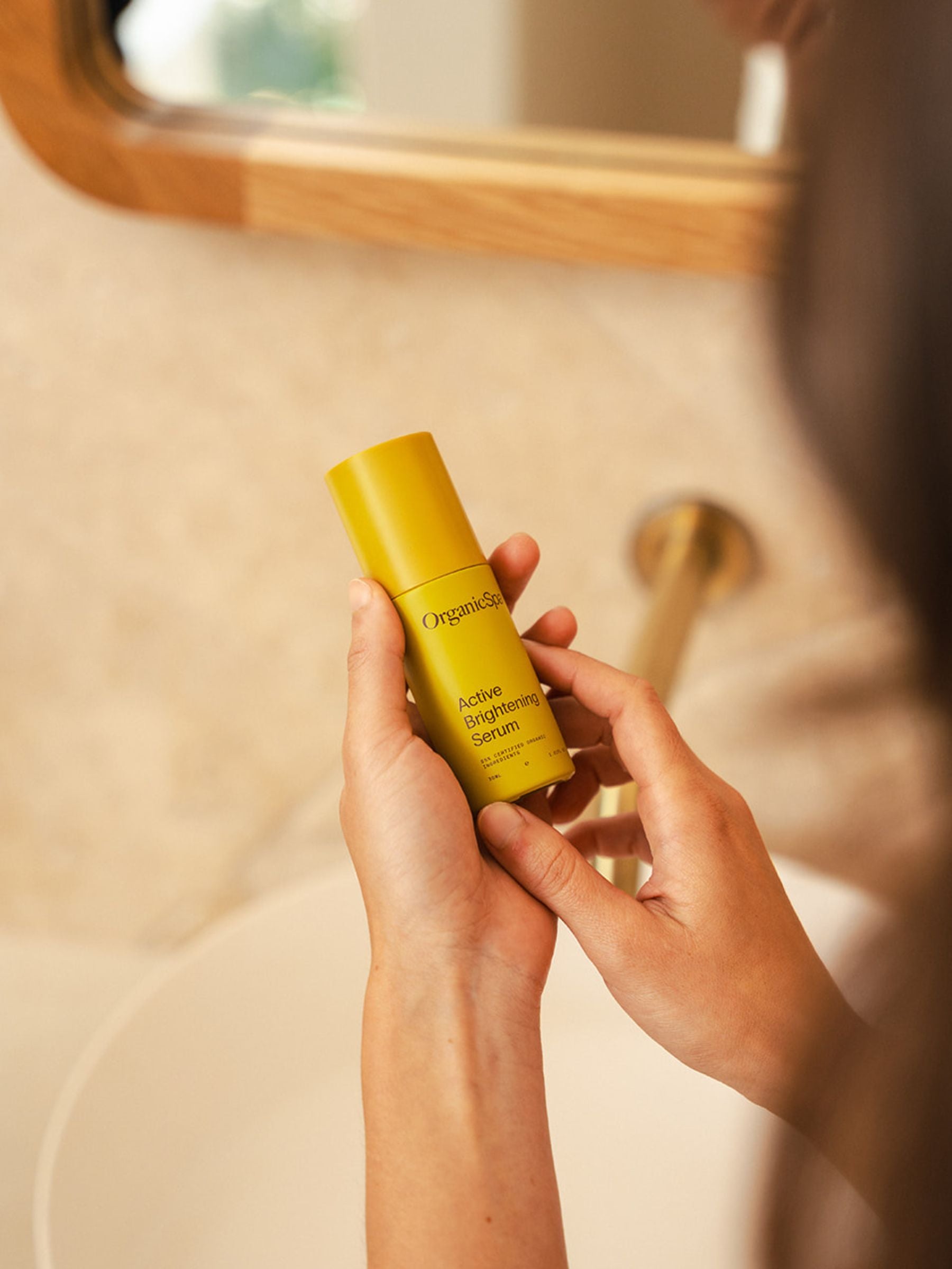 Person holding a bottle of Organicsceuticals Active Brightening Serum in both hands at a bathroom basin, with the mirror and tap softly blurred in the background.