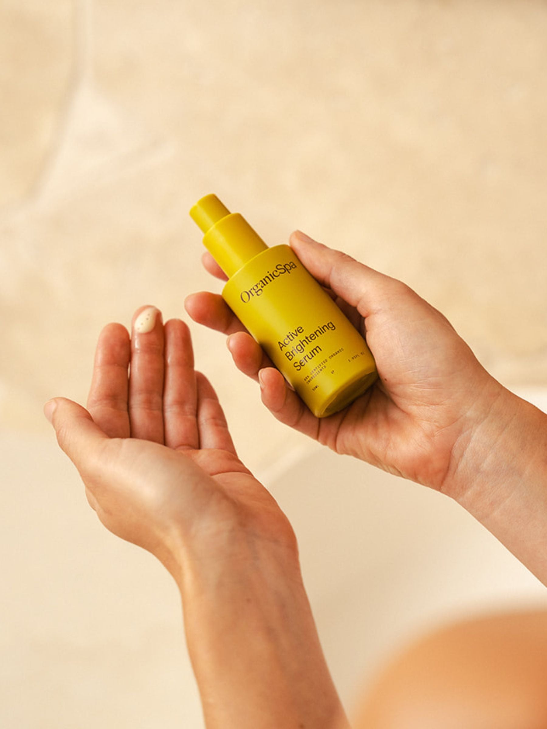 Close-up of hands dispensing Organicsceuticals Active Brightening Serum into the palm, showing the creamy dotted texture of the product against a warm bathroom background.