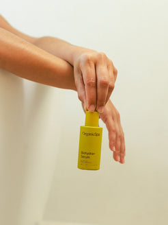 Close-up of an arm resting on a bath edge, hand loosely holding a bright yellow bottle of Organicspa Organicsceuticals Biohydra+ Serum against a soft, neutral background.