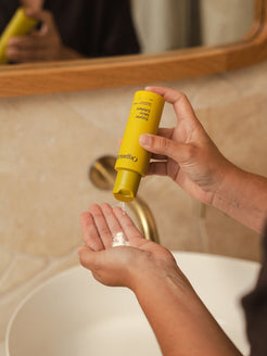Person pouring Organicspa Organicsceuticals Enzyme Micro Exfoliant powder from a yellow bottle into their palm in front of a bathroom mirror.