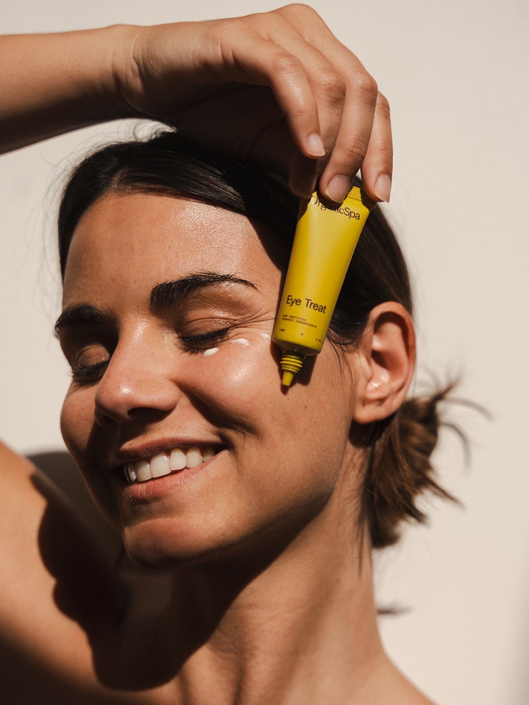 Smiling woman applying Organicspa Organicsceuticals Eye Treat under her eye with the yellow tube, showing the certified organic eye cream in use.