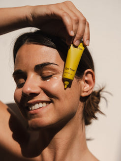 Smiling woman applying Organicspa Organicsceuticals Eye Treat under her eye with the yellow tube, showing the certified organic eye cream in use.