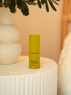 Bottle of Organicspa Organicsceuticals Pro AHA Treatment on a bedside table beside a ceramic vase and greenery, showing the certified organic nightly resurfacing treatment in a calm bedroom setting.
