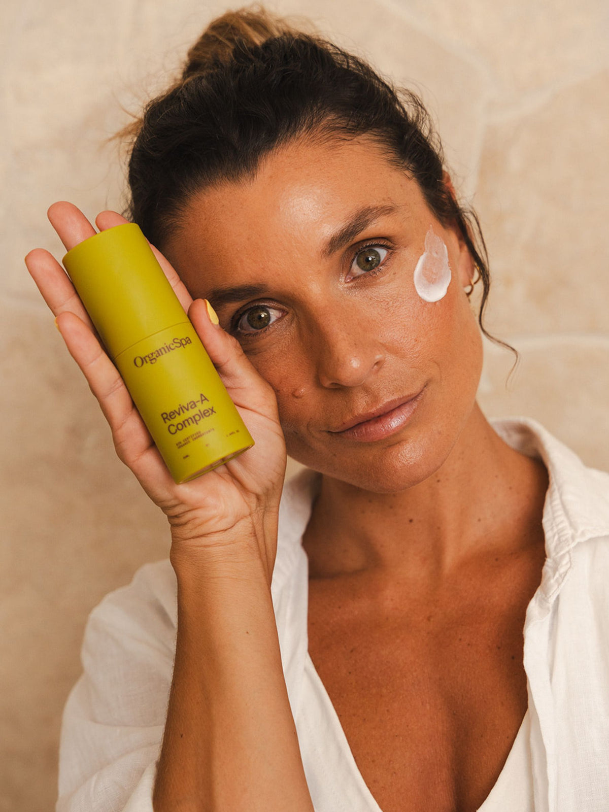 Woman holding OrganicSpa Reviva-A Complex beside her face, with a dab of the lotion on her cheek.