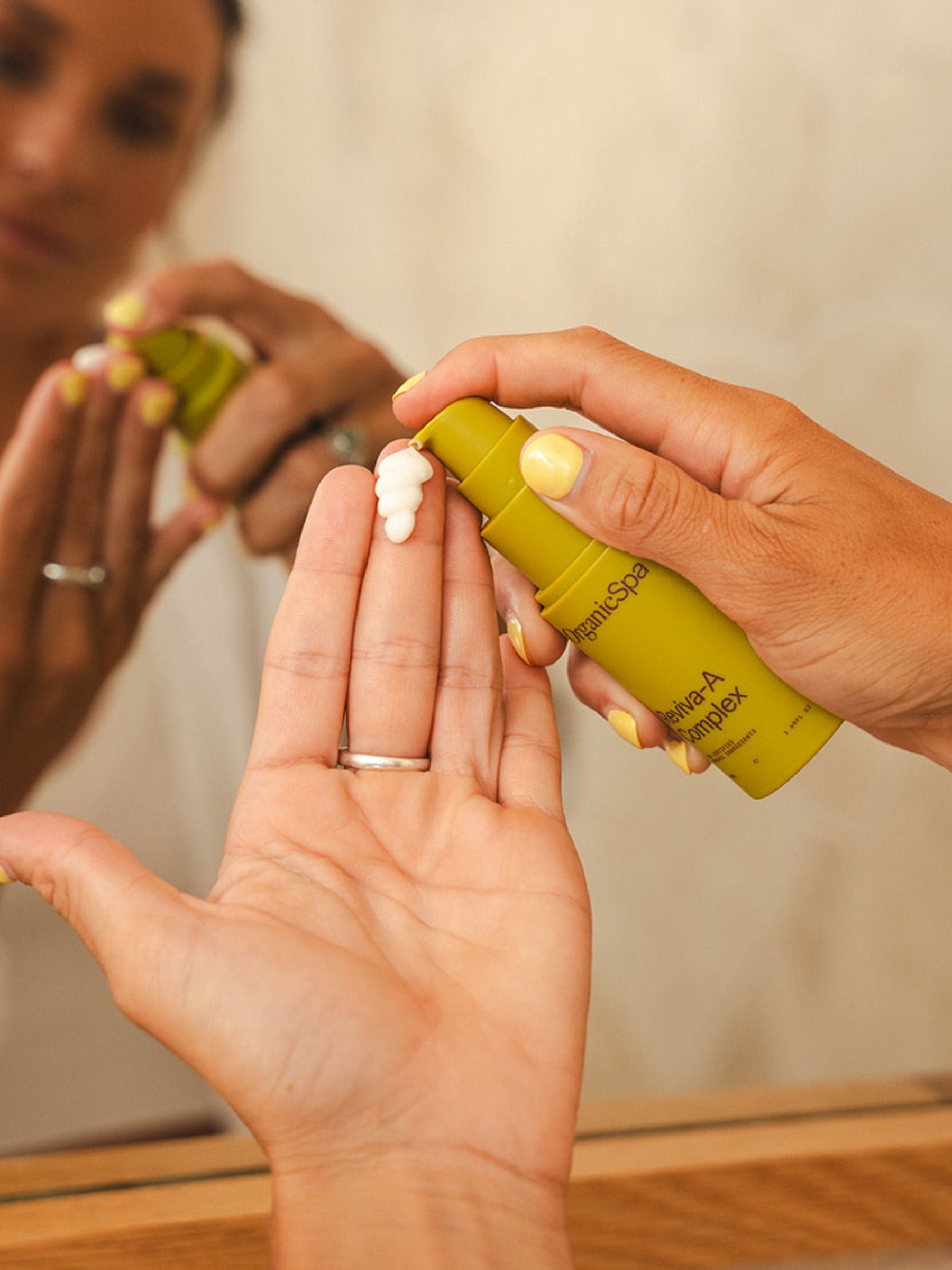 Hands dispensing Organicspa Organicsceuticals Reviva-A Complex serum onto fingertips in front of a bathroom mirror.