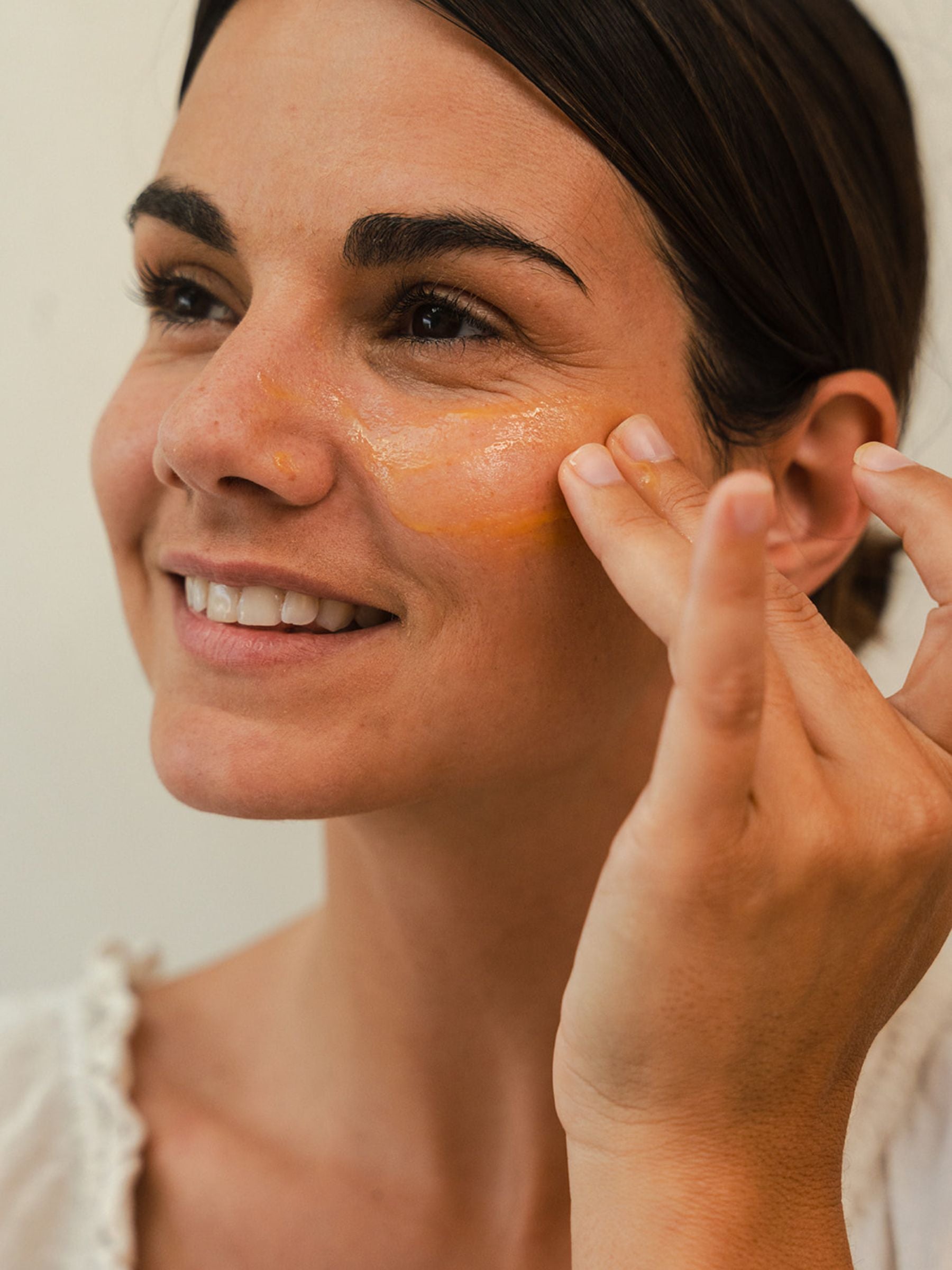 Woman smiling as she applies a thin layer of bright orange Organicspa Organicsceuticals Vita-A+CE Complex serum under her eye and across the upper cheek.