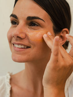 Woman smiling as she applies a thin layer of bright orange Organicspa Organicsceuticals Vita-A+CE Complex serum under her eye and across the upper cheek.