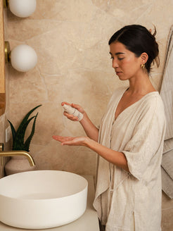 Woman in a robe standing at a bathroom basin, dispensing Organicspa Vitamin C Serum into her palm as part of her daily skincare routine.