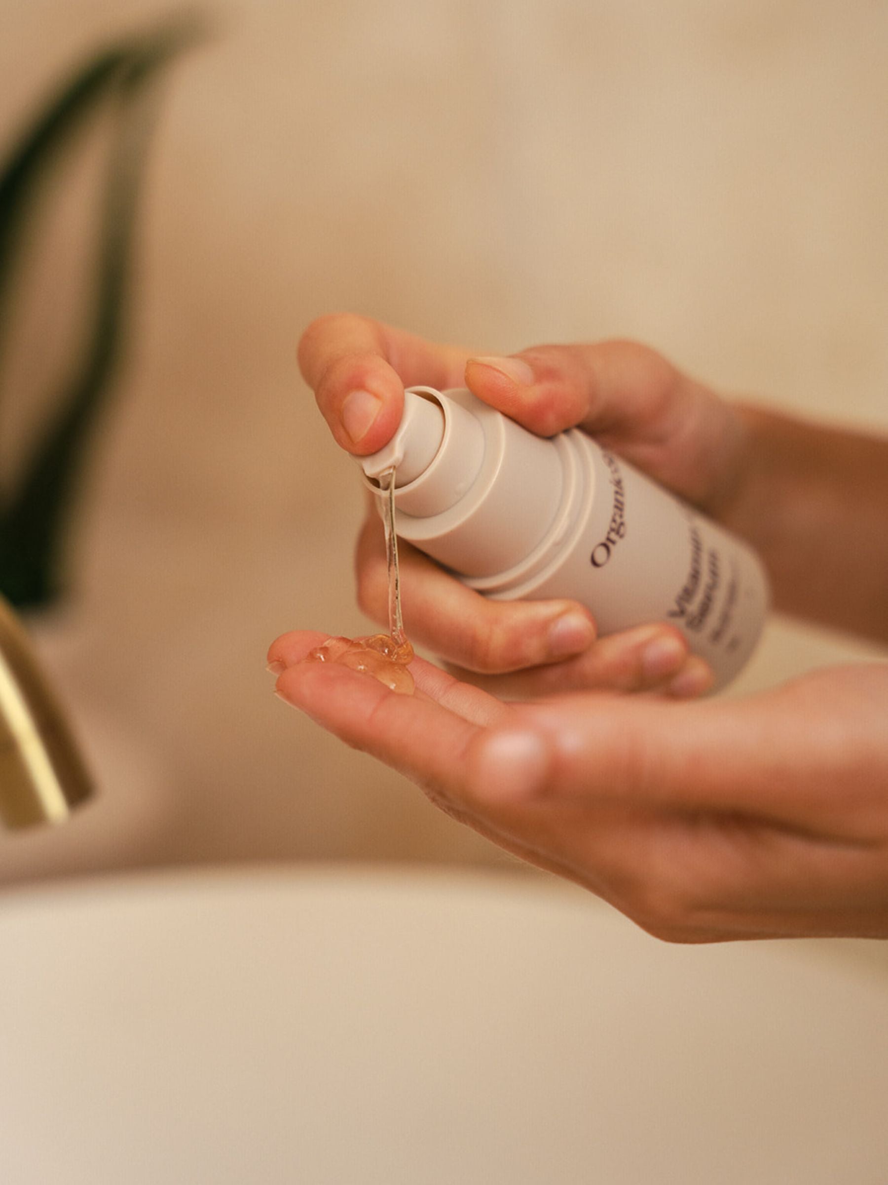 Close-up of a hand pumping a golden drop of Organicspa Vitamin C Serum into the palm, showing the lightweight gel texture beside a bathroom sink.