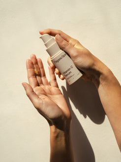 Hands holding and dispensing Organicspa Vitamin C Serum in natural sunlight against a soft neutral wall, highlighting the brightening face serum.