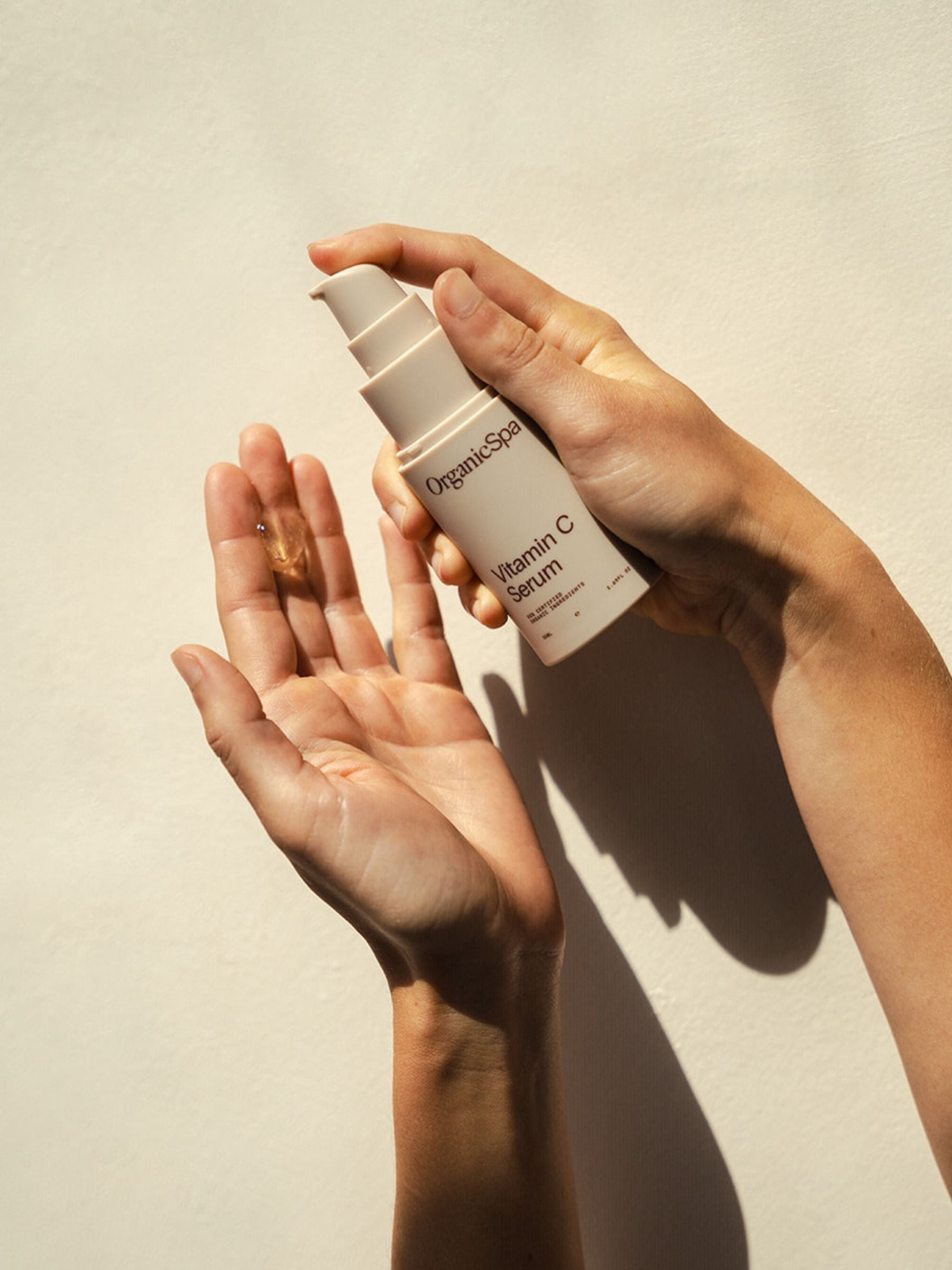 Hands holding and dispensing Organicspa Vitamin C Serum in natural sunlight against a soft neutral wall, highlighting the brightening face serum.