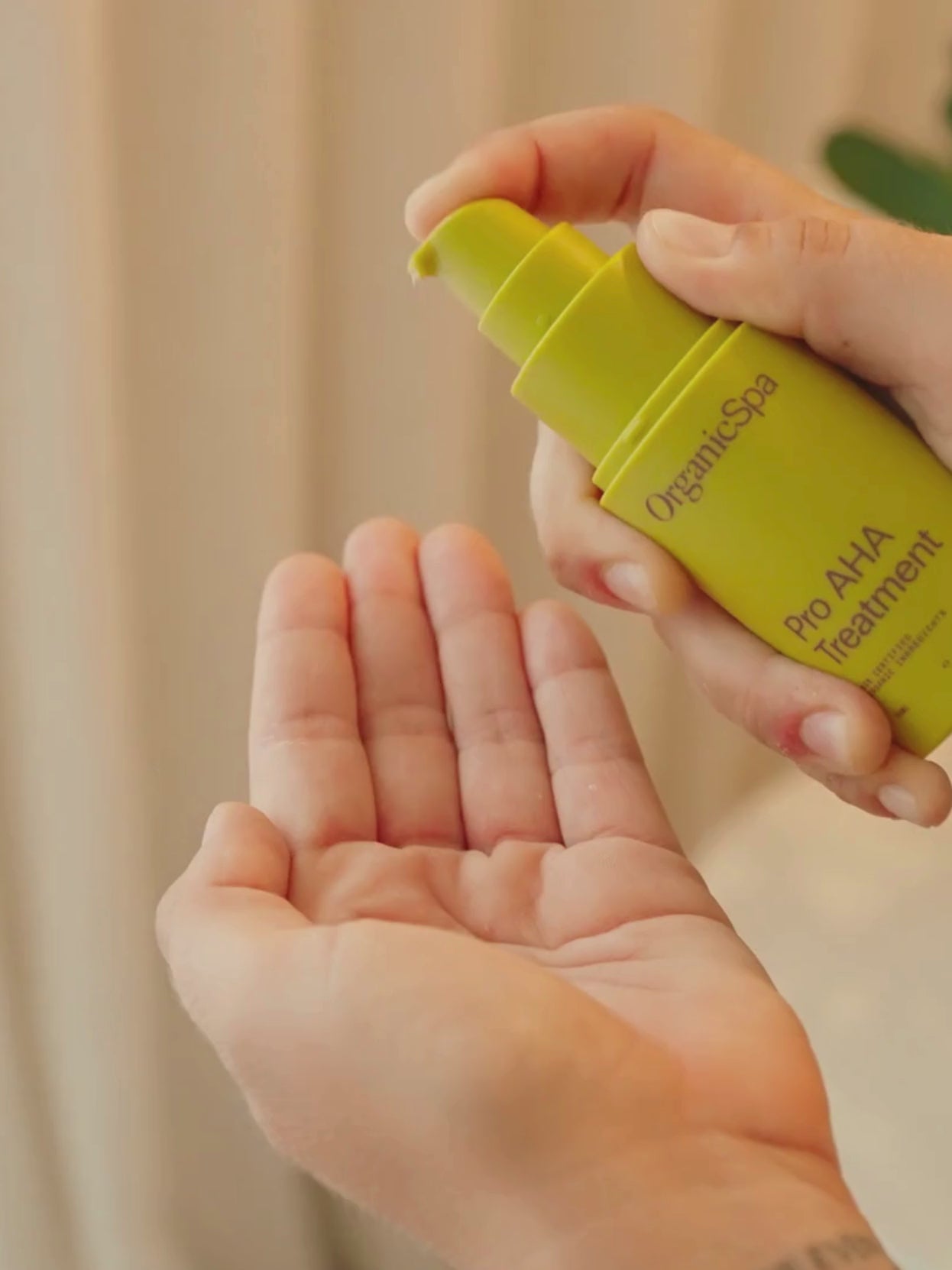 A woman in a sage linen robe dispenses OrganicSpa Pro AHA Treatment into her palm, then massages it over her cheeks in a warm, softly lit room with a native botanical arrangement nearby.