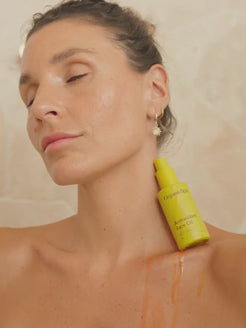A woman tilts her head back with eyes closed as OrganicSpa Antioxidant Face Oil is poured along her collarbone, the rich amber oil catching the warm light against her skin. Certified Organic Skincare.