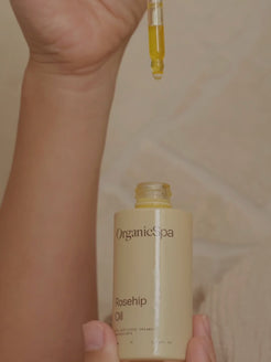 A dropper dispenses a golden drop of OrganicSpa Rosehip Oil onto the inner forearm, the cream bottle held in hand against a soft neutral background.