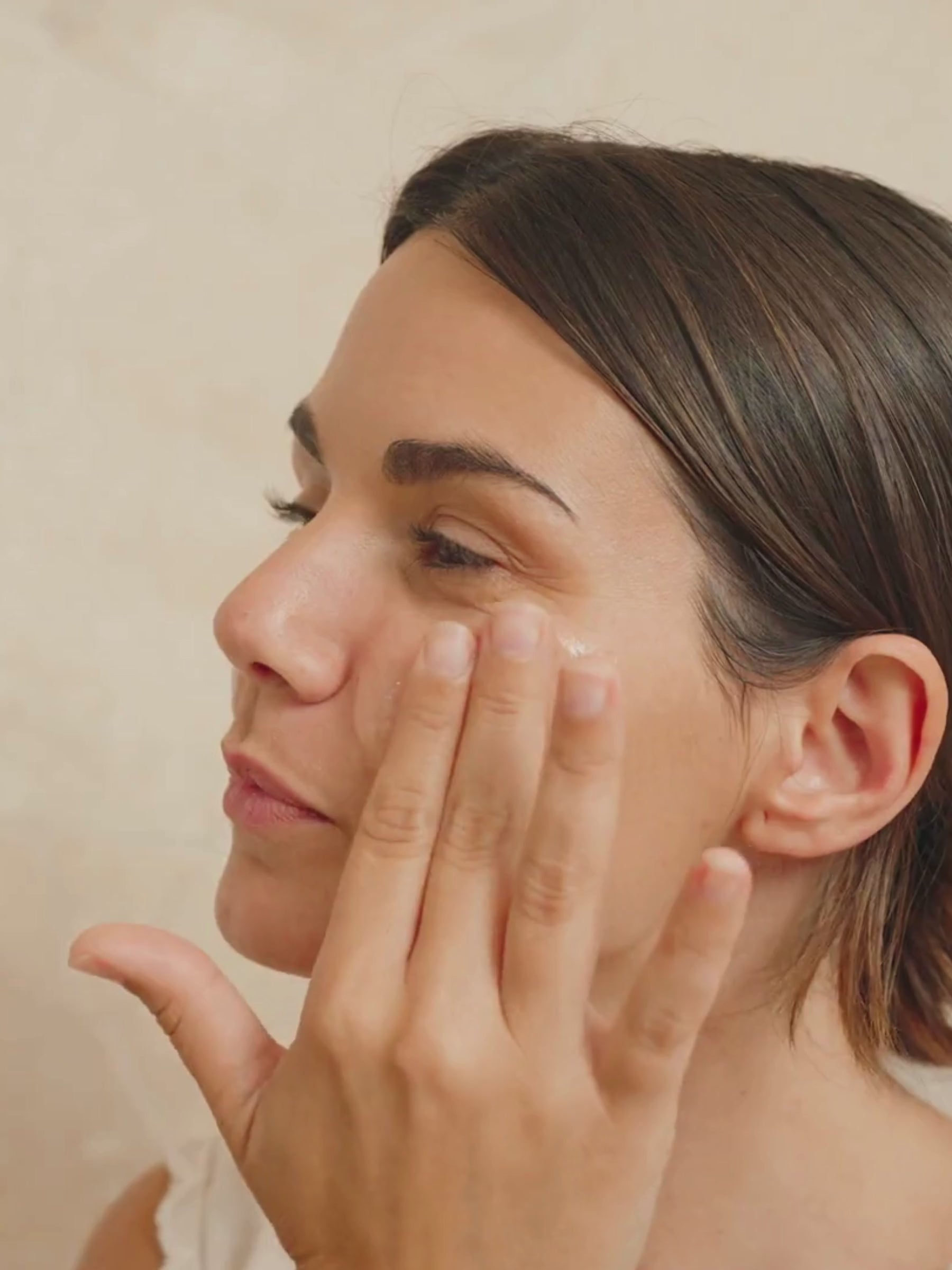 A woman gently presses Organicsceuticals Biohydra+ Serum into the skin around her eye and cheek in a soft side profile, before the video moves into a close-up of the serum absorbing into the skin, visibly dewy. Warm neutral background, slow and unhurried movement. Certified Organic Skincare.