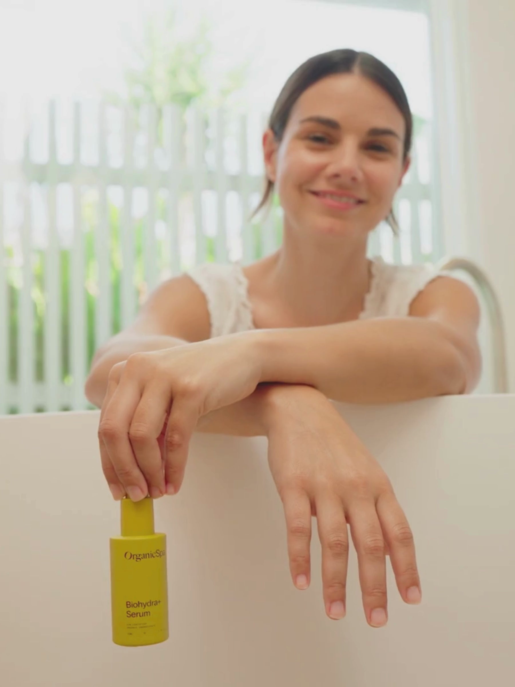A smiling woman leans over the edge of a freestanding bath, dangling the OrganicSpa Biohydra+ Serum from her fingertips, with green foliage and soft natural light behind her. The video moves into a close-up of the bottle held upright, label fully visible. Certified Organic Skincare.