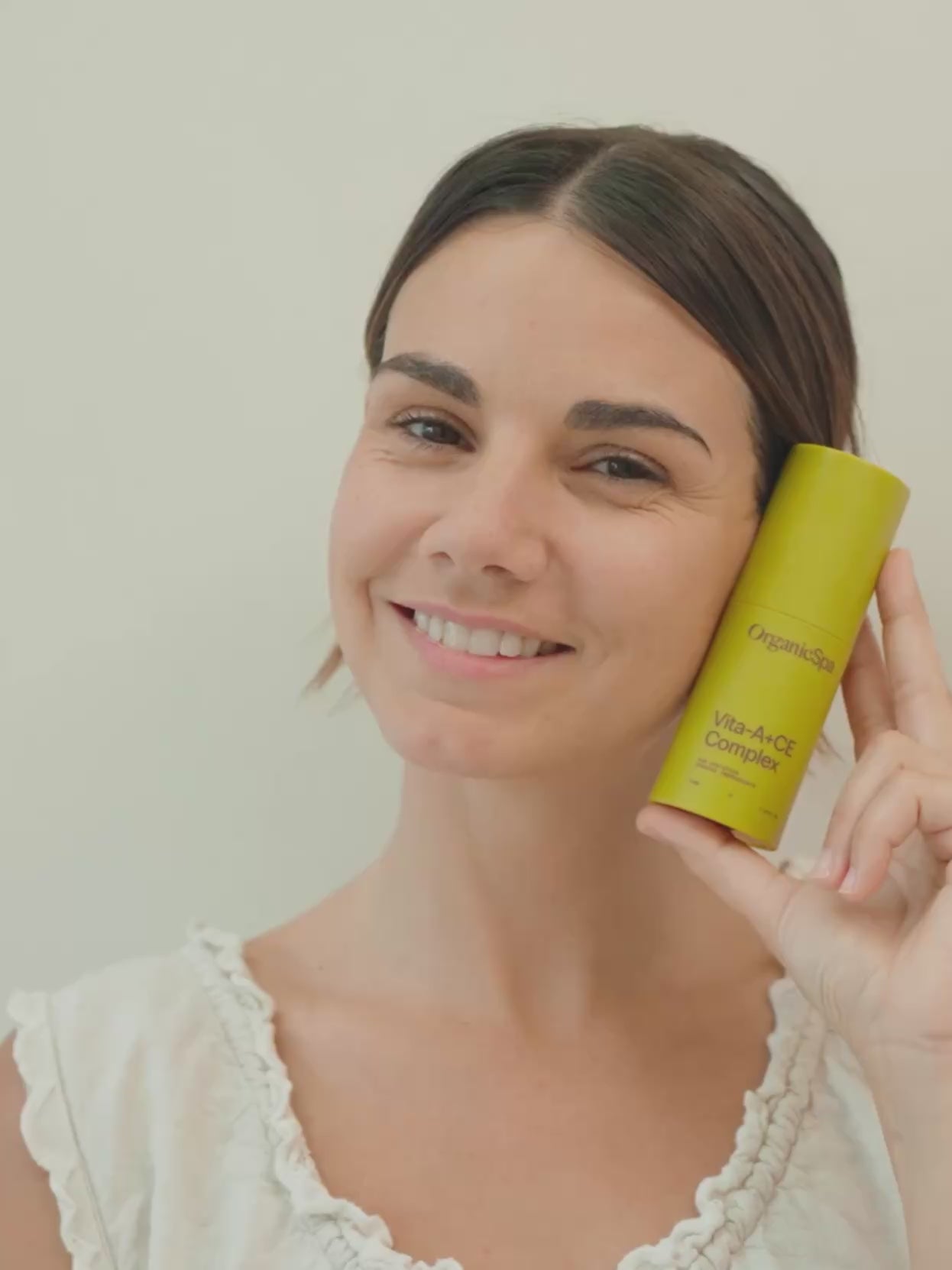 A smiling woman holds the OrganicSpa Vita-A+CE Complex beside her cheek, then the video moves into a close-up of the product resting against her skin, label clearly visible. Soft natural light against a clean white background. Certified Organic Skincare.