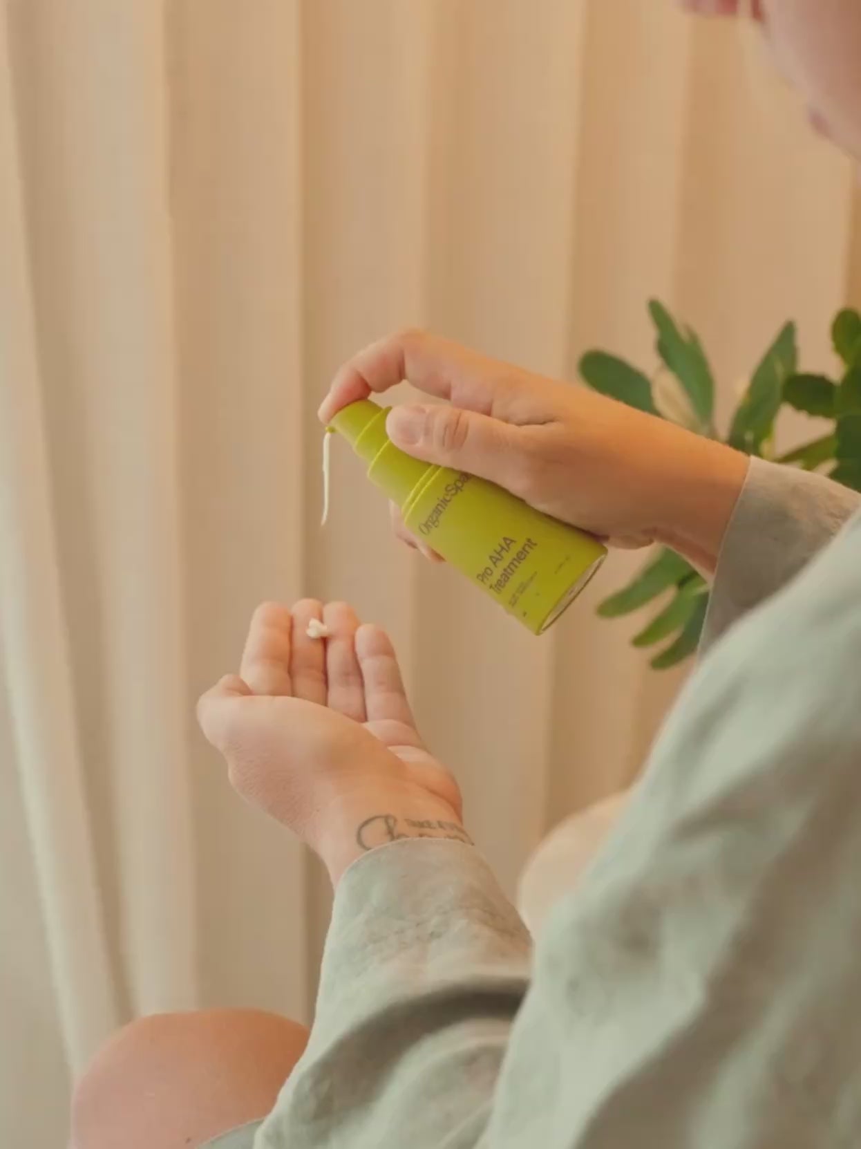 A woman pumps OrganicSpa Pro AHA Treatment into her palm, then gently presses and massages the formula into her face. She wears a soft sage linen robe in a warm, naturally lit room styled with a woven pendant light and white ceramic vase with greenery. Certified Organic Skincare.
