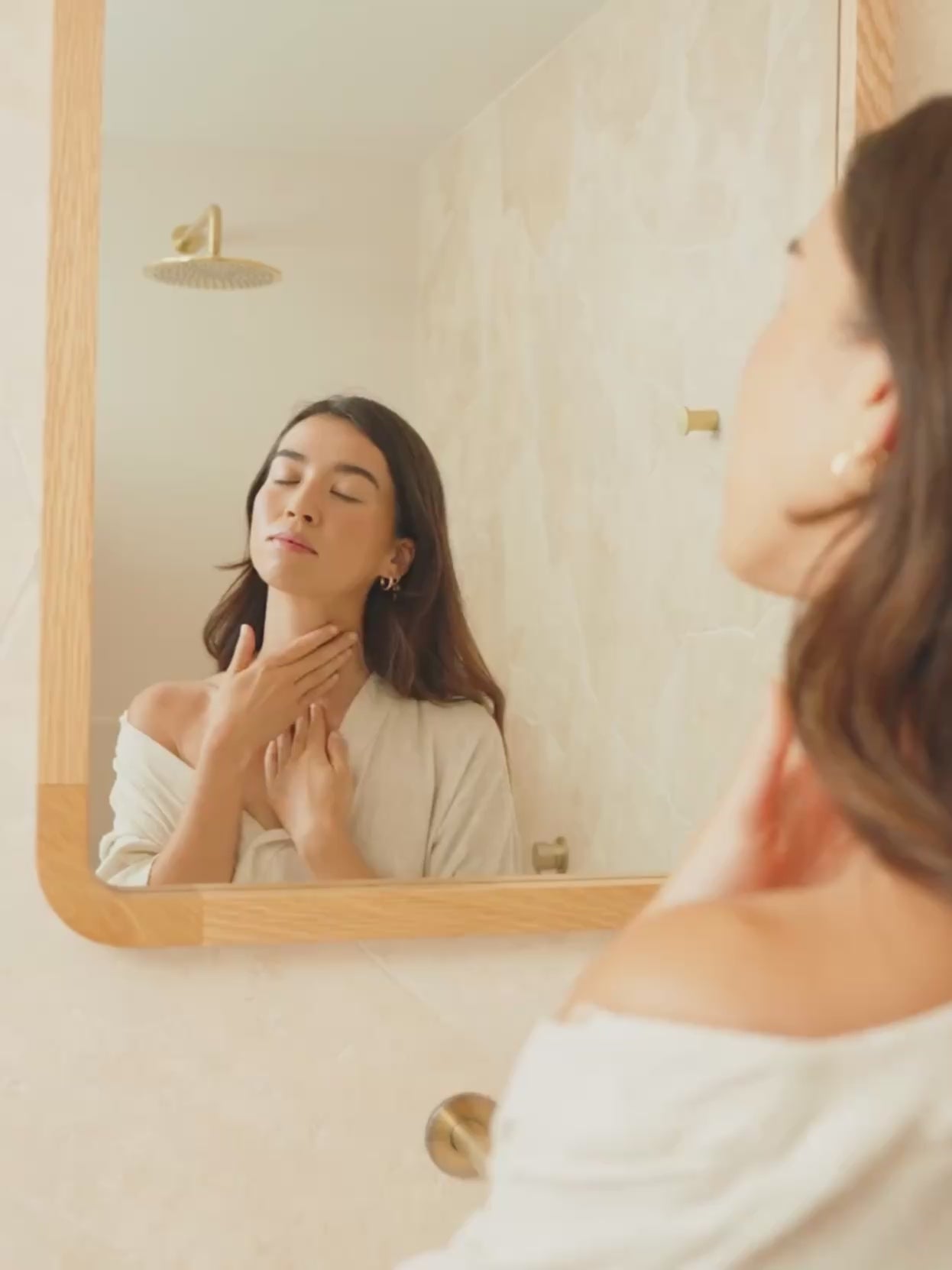 A woman gently massages product into her neck and décolletage, eyes closed, first reflected in a warm timber-framed bathroom mirror, then shown in close-up against a soft textured wall. Gold hardware and natural light throughout. Certified Organic Skincare.