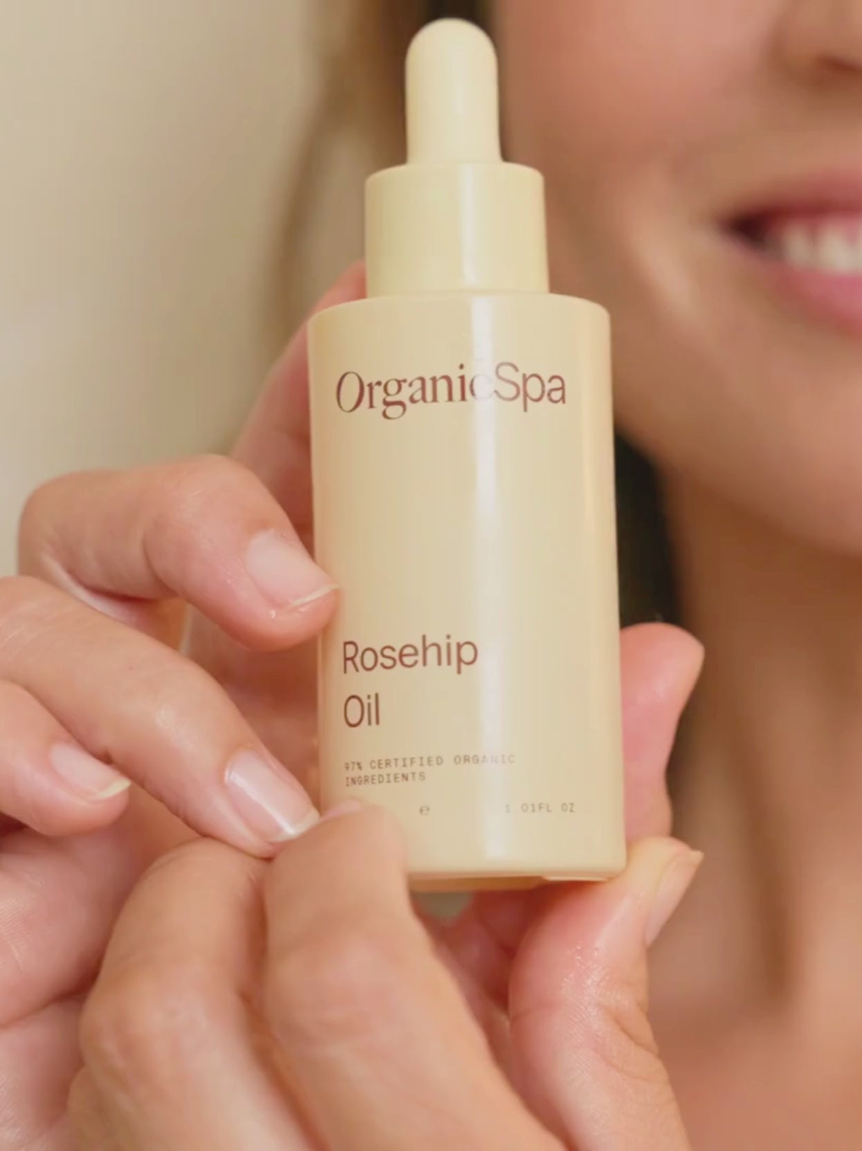 A smiling woman holds up a bottle of OrganicSpa Rosehip Oil to the camera in soft, natural light.
Longer description: Close-up of a smiling woman presenting the OrganicSpa Rosehip Oil dropper bottle toward the camera in a warmly lit room.