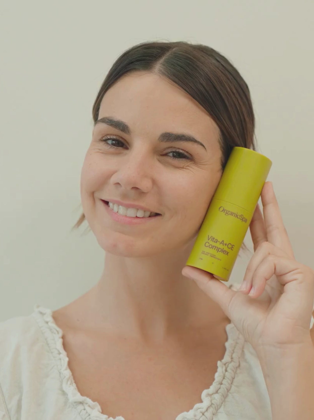 A smiling woman holds a bottle of OrganicSpa Vita-A+CE Complex beside her face in soft, natural light.
Longer description: Close-up of a smiling woman presenting the OrganicSpa Vita-A+CE Complex bottle next to her face in a warmly lit room.