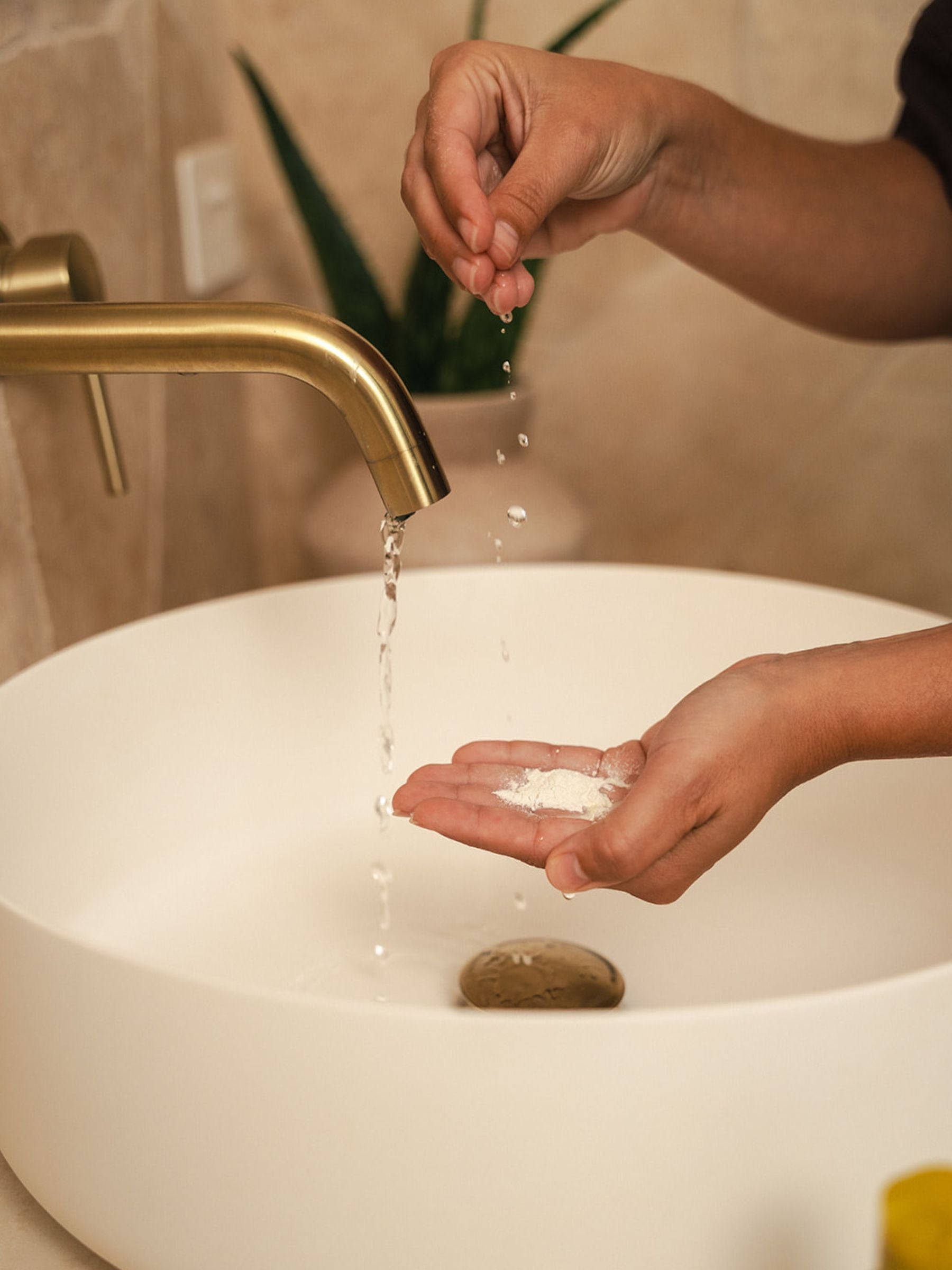 Hands cupping Organicspa Organicsceuticals Enzyme Micro Exfoliant powder under a running tap, activating the Certified Organic enzyme exfoliant at the bathroom sink.