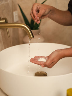 Hands cupping Organicspa Organicsceuticals Enzyme Micro Exfoliant powder under a running tap, activating the Certified Organic enzyme exfoliant at the bathroom sink.